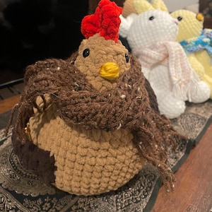 May include: A brown crocheted chicken wearing a brown scarf with a red comb and yellow beak. The chicken is sitting on a patterned surface.