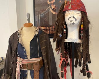 May include: A brown and black pirate coat with a blue vest and a brown leather belt. A red bandana with gold beads and a long brown wig with red and pink accents are on a stand behind the coat. The poster behind the coat says "Pirates of the Caribbean: Curse of the Black Pearl."