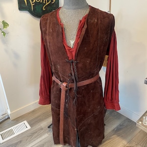 May include: A brown suede vest with a matching belt over a red long-sleeved shirt. The vest has a braided closure and a split front. A silver pendant hangs from the neckline. The Prancing Pony sign is visible in the background.