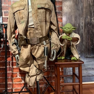 May include: A brown and tan jumpsuit with a belt and a lightsaber. A green Yoda figurine is standing on a wooden stool.