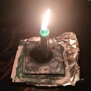 May include: A lit green candle stands upright on a square, layered base with a marbled green and white surface. The base is partially wrapped in crinkled silver aluminum foil, with melted green wax pooling around the candle's base on the square.