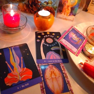 May include: A collection of oracle cards featuring colourful illustrations and text including Be Still, Look to the Stars, Support, Intention, and Animals. The cards are arranged near a red candle in a glass holder and a tea light candle in an apple.