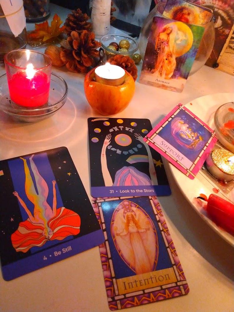May include: A collection of oracle cards featuring colourful illustrations and text including Be Still, Look to the Stars, Support, Intention, and Animals. The cards are arranged near a red candle in a glass holder and a tea light candle in an apple.