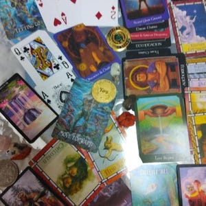 May include: A collection of tarot cards, playing cards, and small metal coins scattered together. The cards feature various colourful illustrations, including figures, landscapes, and text like 'Move Forward', 'Children', and 'Love Begins'.