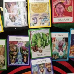 May include: A collection of rectangular oracle cards with colourful illustrations and white borders. The cards feature various titles including Dissipating, Abundance, Soulmate, Facade, Gossip, Faith, Trickery, Vanity, Workaholic, and Balance.