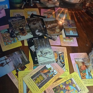 May include: A collection of tarot cards spread across a surface, featuring various illustrations including a wolf, swords, and pentacles. The cards display text such as Girl Fight, Fake, Queen of Wands, and King of Wands, with colourful imagery.