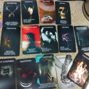May include: A collection of rectangular oracle cards featuring dark, evocative imagery and text labels. Each card displays a specific theme such as Stubborn, Sensitive, Jealous, Wicked, Predator, Narrow Mind, Overthinker, Abusive, Emotionally Unavailable, Self-Sabotage, Trusting, and Cheater, accompanied by descriptive keywords.