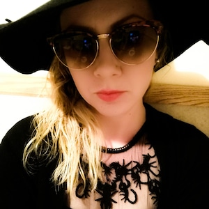 Puede incluir: Un sombrero negro de ala ancha, gafas de sol con patrón de carey y monturas doradas, y un cárdigan negro con un panel transparente en el cuello adornado con motivos de arañas bordados en negro y un borde de cuentas.
