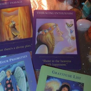 May include: Four rectangular oracle cards with purple and green borders. One card shows a person with a flower crown and butterflies, labelled Embracing Enthusiasm. Another shows a desert scene, labelled Desert Passage. Others show angels.
