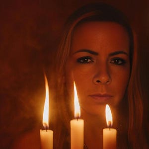 May include: Three white pillar candles with bright, flickering orange flames positioned in the foreground. The light from the candles illuminates the face of a person with long hair, creating a warm, glowing atmosphere in a dimly lit environment.