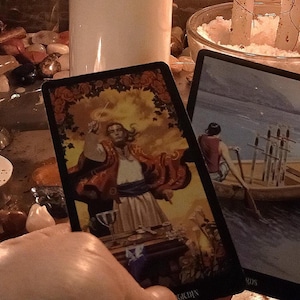 May include: Two rectangular tarot cards featuring detailed illustrations. The left card depicts a figure in a red robe standing at a table, while the right card shows a person rowing a boat containing six upright swords across calm water.