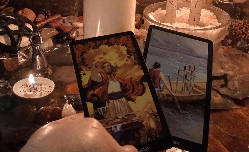 May include: Two rectangular tarot cards featuring detailed illustrations. The left card depicts a figure in a red robe standing at a table, while the right card shows a person rowing a boat containing six upright swords across calm water.