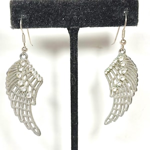 K&ouml;nnte beinhalten: Ein Paar silberfarbene Fl&uuml;gelohrringe mit klaren Kristallakzenten. Die Ohrringe werden auf einem schwarzen Schmuckst&auml;nder vor wei&szlig;em Hintergrund pr&auml;sentiert. Die Fl&uuml;gel haben ein detailliertes, offenes Design.