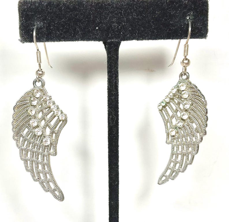K&ouml;nnte beinhalten: Ein Paar silberfarbene Fl&uuml;gelohrringe mit klaren Kristallakzenten. Die Ohrringe werden auf einem schwarzen Schmuckst&auml;nder vor wei&szlig;em Hintergrund pr&auml;sentiert. Die Fl&uuml;gel haben ein detailliertes, offenes Design.