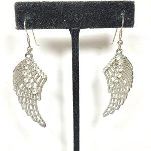 K&ouml;nnte beinhalten: Ein Paar silberfarbene Ohrringe in Fl&uuml;gelform. Jeder Ohrring hat ein Fl&uuml;geldesign mit filigranen Details und ist mit klaren, funkelnden Kristallen verziert. Die Ohrringe h&auml;ngen an einem einfachen Haken.