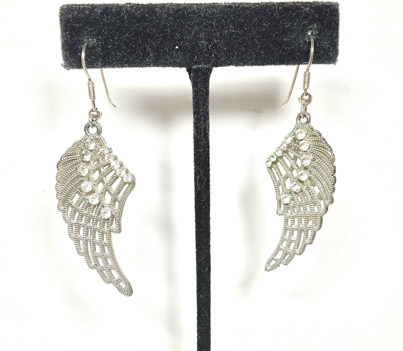 K&ouml;nnte beinhalten: Ein Paar silberfarbene Ohrringe in Fl&uuml;gelform. Jeder Ohrring hat ein Fl&uuml;geldesign mit filigranen Details und ist mit klaren, funkelnden Kristallen verziert. Die Ohrringe h&auml;ngen an einem einfachen Haken.