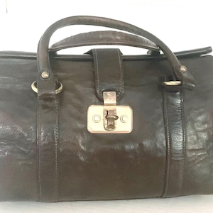 May include: A dark brown leather handbag with a smooth, slightly distressed texture. It features a single top handle, a central vertical strap with a rectangular metal clasp closure, and silver-toned hardware rings connecting the handle to the bag.