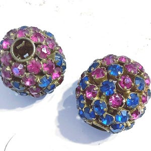 May include: Two vintage, round earrings. Each earring is covered in small, faceted, pink and blue rhinestones. The earrings have a gold-toned metal base. One earring has a visible post.