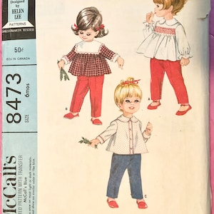 May include: Vintage sewing pattern for toddler's clothing. The pattern includes three tops with transfer for smocking and pants. The pattern is for size 6 months and is printed on blue paper.