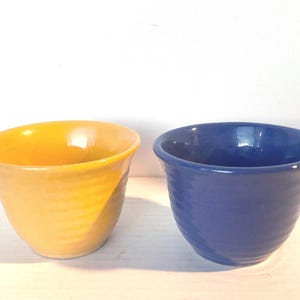 May include: Two small ceramic bowls with a smooth, glossy finish. One bowl is a vibrant, sunny yellow, and the other is a deep, rich royal blue. Both bowls feature a subtle, horizontal ribbed texture along their rounded, tapered sides.