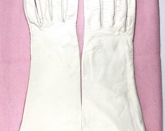 May include: A pair of white leather gloves with long sleeves, laid out on a pink surface. The gloves have visible stitching along the fingers and seams. The leather appears smooth and the gloves are clean.
