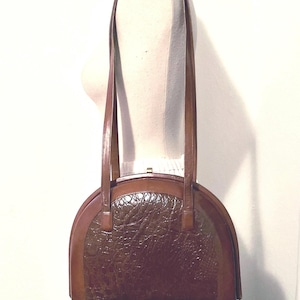 May include: A vintage brown handbag with a semi-circular shape. The bag features a textured, crocodile-like pattern on the front panel, framed by smooth, darker brown leather. It has two long, slender handles and a gold-toned clasp.
