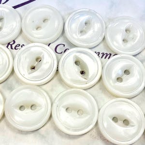 May include: A collection of fourteen round, white buttons arranged in three rows. Each button features a smooth, glossy finish with a slightly recessed center containing two holes for attachment. The buttons have a uniform circular shape.