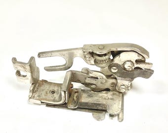 May include: A vintage, silver-toned sewing machine attachment. The device has multiple levers, gears, and a hinged design. It is designed to be attached to a sewing machine. The attachment is made of metal and has a worn appearance.