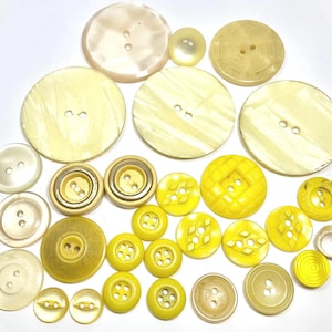 May include: A collection of various yellow and cream-colored buttons. The buttons vary in size and design, including round, flat, and textured styles. Some have two or four holes, while others have decorative patterns. The buttons are arranged on a white surface.
