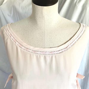 May include: Pale pink sleeveless top with a scoop neckline trimmed with delicate lace. The top has a soft, flowing fabric and is displayed on a mannequin. Pink ribbons are attached at the sides. A vintage-inspired garment.