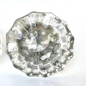 May include: Clear, faceted glass cabinet knob with a decorative, textured design. The knob has a round shape with a scalloped edge. The glass is transparent, allowing light to pass through.