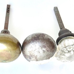 May include: A collection of antique door knobs with metal stems. The knobs include a brass sphere, a tarnished silver sphere, and a clear, faceted glass knob. The stems are aged and show signs of wear.