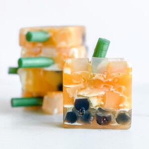 May include: A stack of handmade soap bars with a green swirl design. The top bar has a green stem sticking out of it. The soap bars are layered in a clear container.