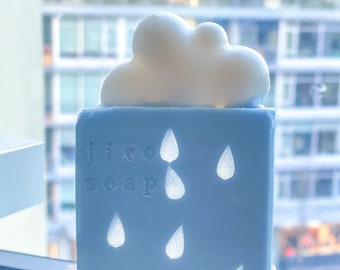 May include: A blue soap bar with a white cloud on top and teardrop cutouts. The soap has the words "jiro soap" etched on the front. The soap is sitting on a wooden soap dish.
