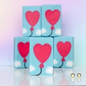 May include: Stack of five blue soap bars with a red heart shaped balloon design. The soap bars are arranged in a pyramid shape. Two gold beads are in the foreground.
