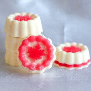 May include: A stack of four white and pink round soaps with a red maple leaf design. The soaps are arranged in a pyramid shape.