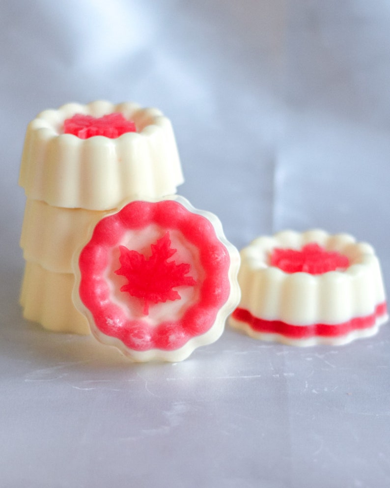 May include: A stack of four white and pink round soaps with a red maple leaf design. The soaps are arranged in a pyramid shape.