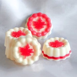 May include: Four white and red scented wax melts shaped like a round cake with a red maple leaf design on top.