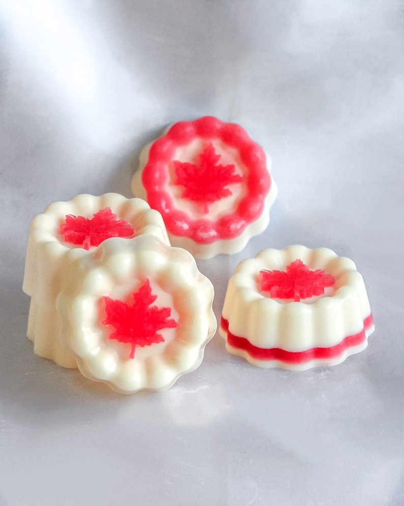 May include: Four white and red scented wax melts shaped like a round cake with a red maple leaf design on top.