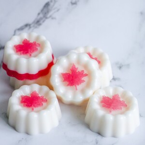 May include: Five white wax melts with a red maple leaf design. The wax melts are shaped like small cakes with a scalloped edge.