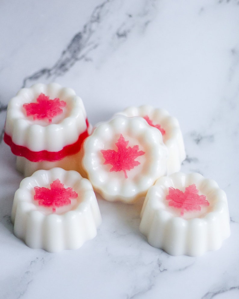 May include: Five white wax melts with a red maple leaf design. The wax melts are shaped like small cakes with a scalloped edge.