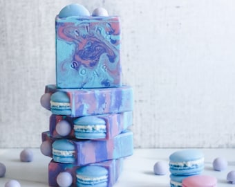 May include: A stack of handmade soaps in shades of blue, purple, and pink, with a marbled effect. The top soap has the text "STAR SOAP". Each soap has a decorative macaron. Small, round, lavender-colored bath bombs and macarons are scattered around.