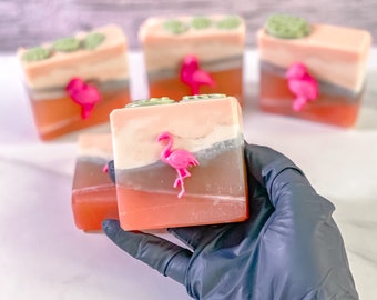 May include: Four handmade soap bars with a tropical theme. Each bar features a pink flamingo and a green leaf design. The soaps are layered in shades of pink, orange, and green.