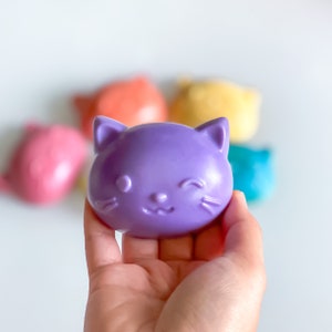 May include: A purple cat-shaped soap bar with a winking face. The soap is in a hand, and there are other colorful soap bars in the background.