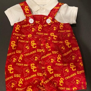 May include: A baby outfit featuring a white polo shirt paired with red shorts and suspenders. The shorts and suspenders are patterned with the text "USC", "TROJANS", and "FIGHT ON!" in yellow.