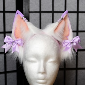 May include: White faux fur cat ears with lavender tips and pink inner ears. The ears are adorned with silver accents and lavender satin bows. The ears are attached to a headband, perfect for cosplay or costume.