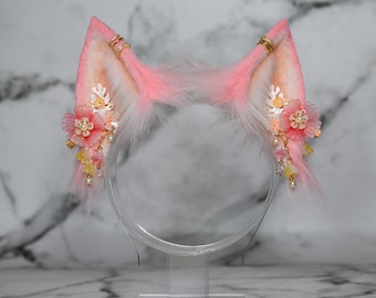 May include: A headband featuring two faux fur cat ears in shades of pink and white. Each ear is decorated with gold-toned metal rings, translucent pink floral embellishments, small white pearls, and dangling beaded accents in yellow and pink.
