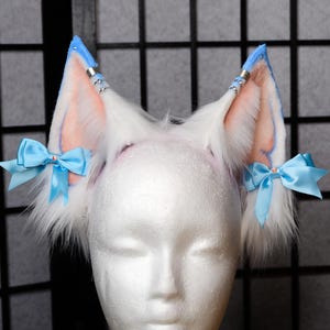 May include: White faux fur fox ears with light blue accents and bows. The ears have a pink interior and are adorned with silver-colored metal accents. The ears are attached to a white headband.