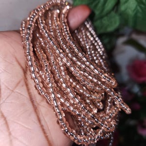May include: A bundle of delicate, rose-gold colored beads. The beads are small, round, and strung together, creating a shimmering effect. The beads are held in a hand, with a blurred background of green leaves and pink flowers.