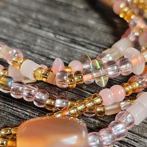 May include: Close-up of several strands of beaded jewelry. The beads are in shades of pink, gold, and clear, with a large, oval, peach-colored bead as a focal point. The jewelry is likely a necklace or bracelet.